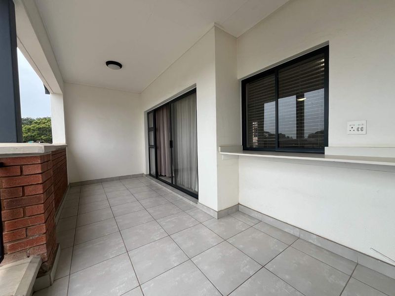 A clean, covered balcony featuring tiled flooring, a brick balustrade, and sliding glass doors providing access to the interior living space.