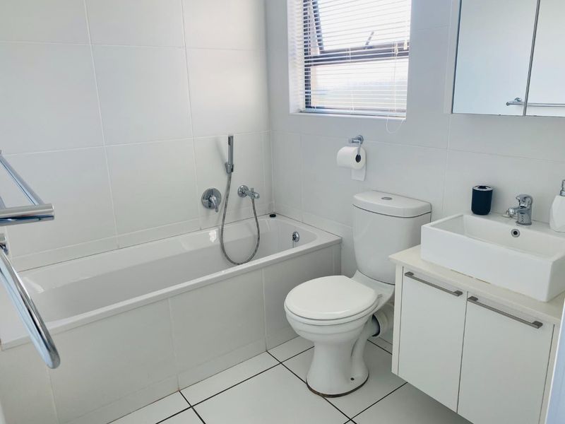 A clean, modern bathroom featuring white wall and floor tiles, a built-in bathtub with handheld shower, a vanity with vessel sink, and a heated towel rail.