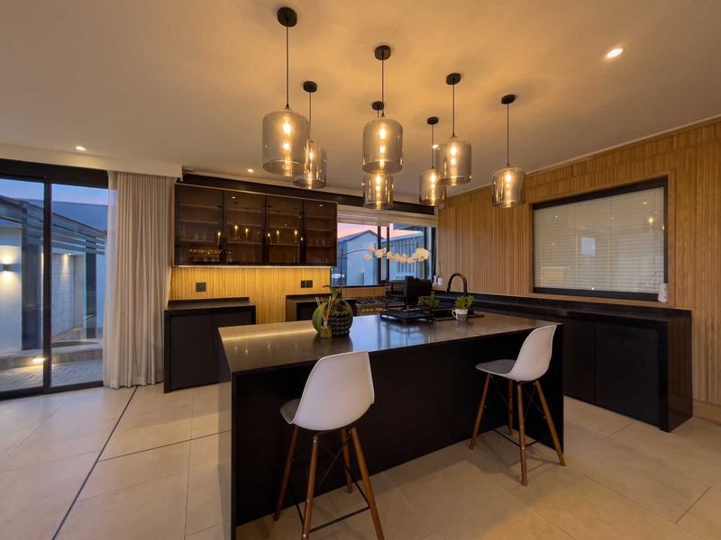 Modern kitchen featuring a large central island, sleek dark cabinetry, stone countertops, and warm wood paneling, complemented by stylish pendant lighting.