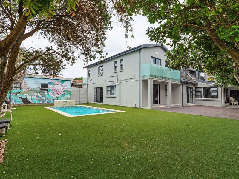 Rear view of the property featuring a swimming pool, paved patio area, glass balcony balustrade, and a well-maintained artificial lawn.