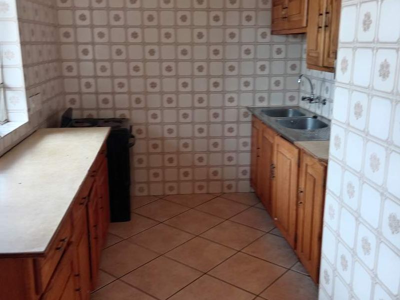 A dated kitchen featuring wooden cabinetry, laminate countertops, a double bowl stainless steel sink, and full-height ceramic wall tiling.