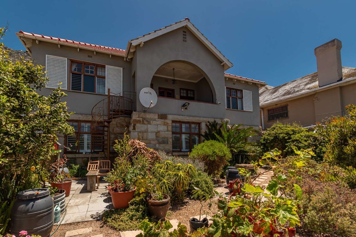 Rear view of a multi-story house featuring stone cladding, a covered balcony, and a spiral staircase leading to a landscaped garden and paved patio area.