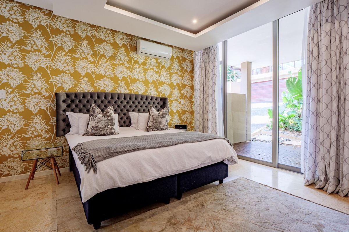 Spacious bedroom featuring a recessed ceiling with downlights, wall-mounted air conditioning unit, and large sliding glass doors opening to a private patio area.