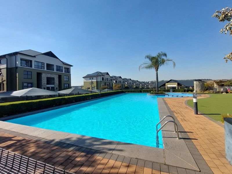 A well-maintained communal swimming pool area featuring paved decking, manicured hedges, and artificial lawn within a modern apartment complex.