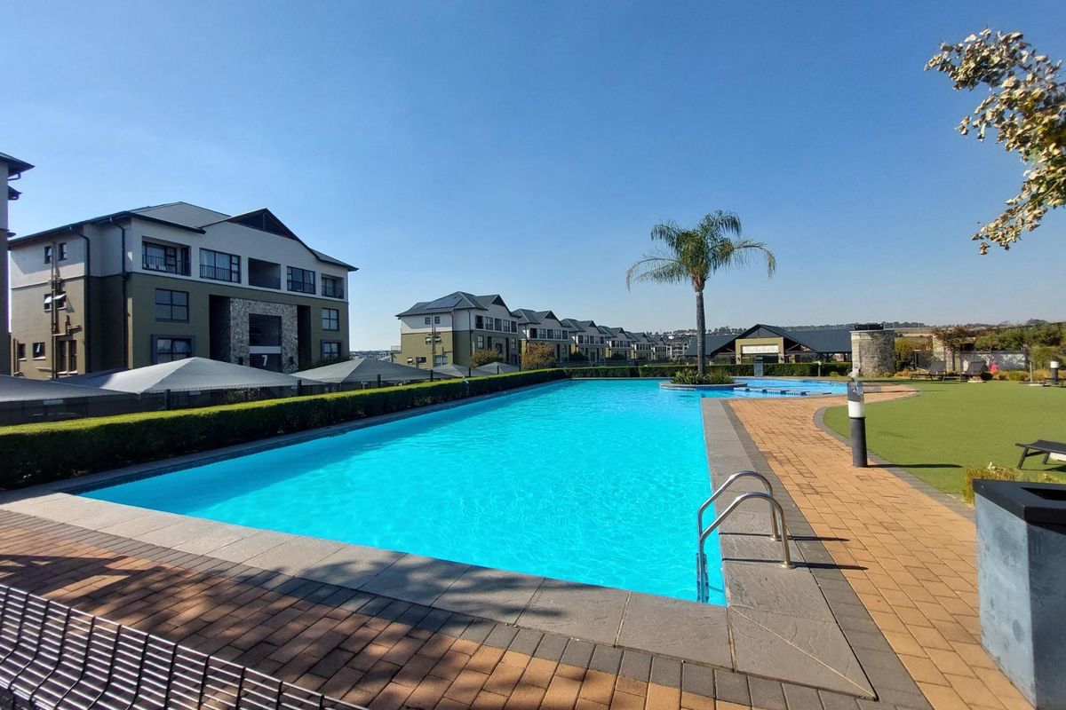 A well-maintained communal swimming pool area featuring paved decking, manicured hedges, and artificial lawn within a modern apartment complex.