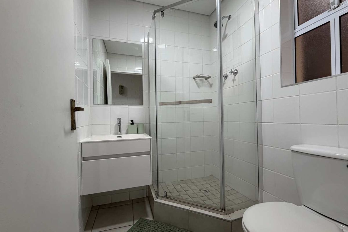 A clean, functional bathroom featuring a corner shower enclosure, wall-mounted vanity with basin, and a standard toilet with white wall tiling.