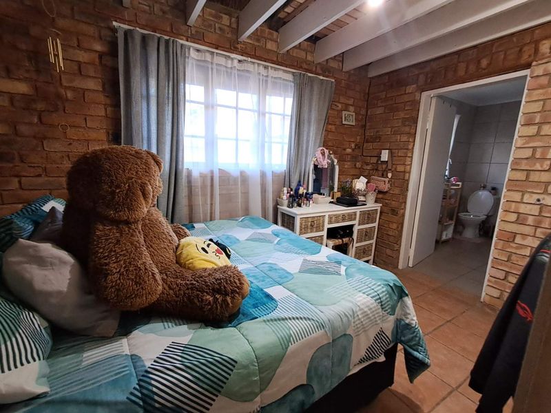 A cozy bedroom featuring rustic exposed brick walls, timber ceiling beams, and tiled flooring, with an adjoining bathroom visible through the doorway.