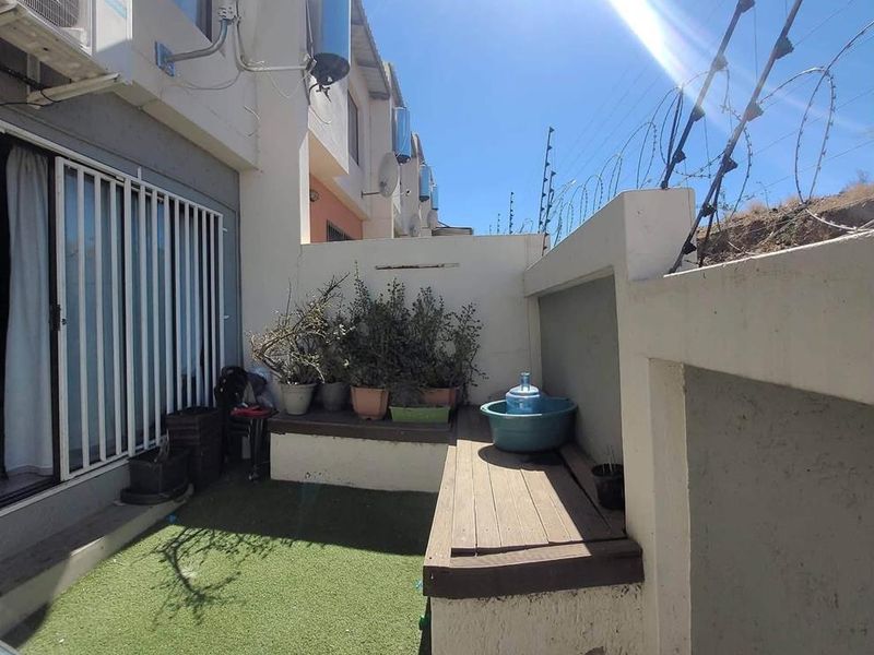 A compact rear courtyard featuring artificial grass, a built-in bench, and security fencing, with visible wear on the boundary wall plaster.