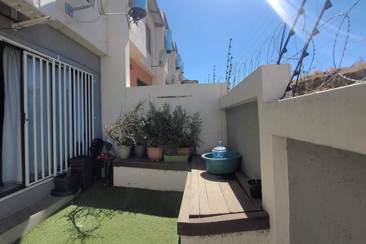 A compact rear courtyard featuring artificial grass, a built-in bench, and security fencing, with visible wear on the boundary wall plaster.