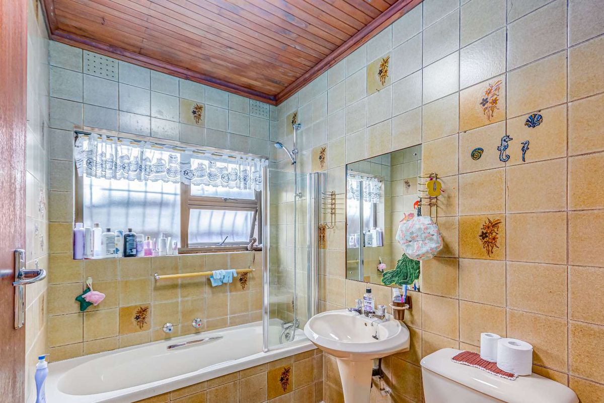 A dated bathroom featuring a bathtub with shower attachment, pedestal sink, toilet, and distinctive timber ceiling with full-height wall tiling.