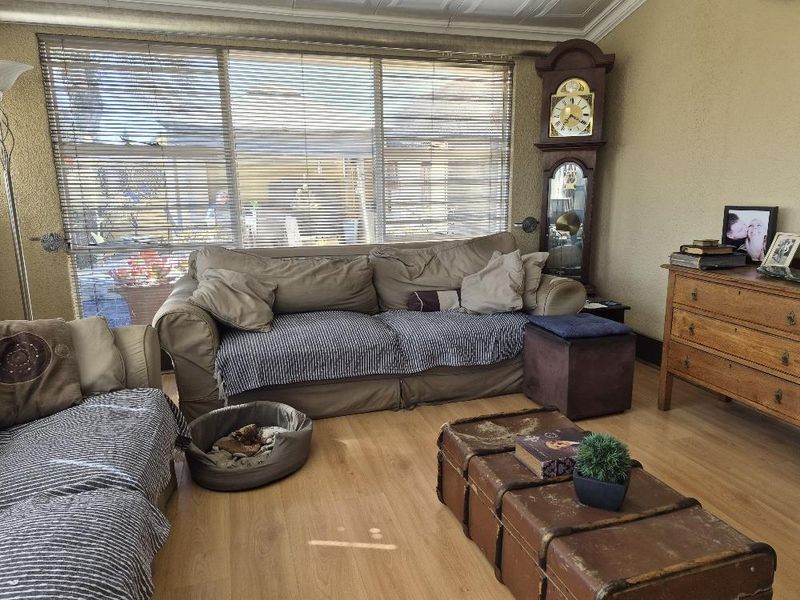 A comfortable living room featuring laminate flooring and large windows with venetian blinds, providing natural light and a view of the outdoor area.