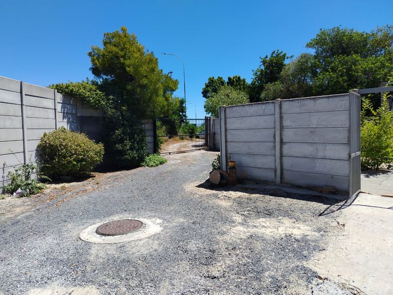 An exterior view of the property entrance featuring a gravel driveway, precast concrete boundary walls, and a metal security gate.