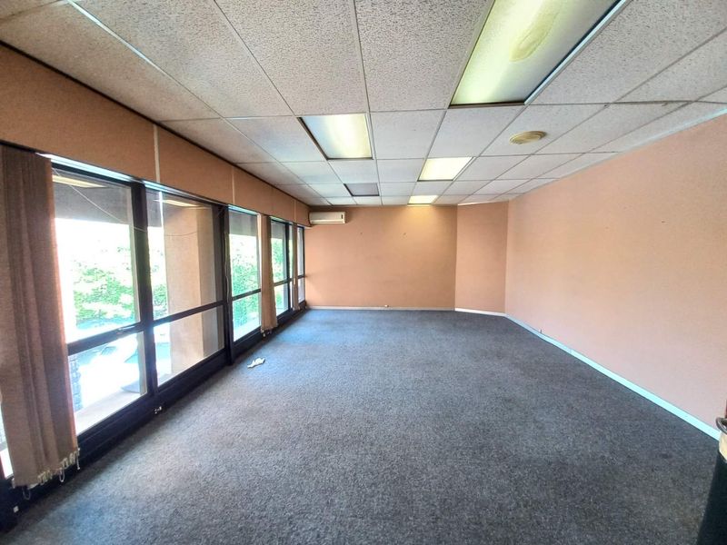 An empty office space featuring large windows, carpeted flooring, a drop ceiling with integrated lighting, and a wall-mounted air conditioning unit.