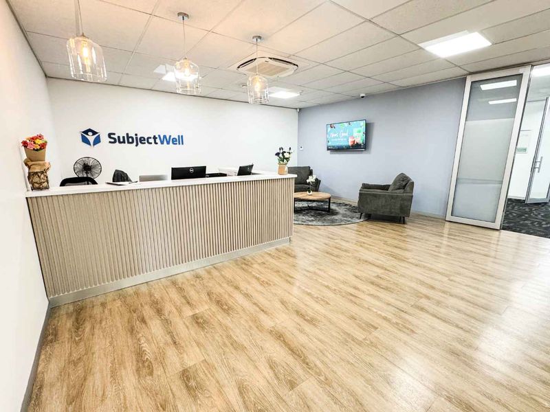 Modern office reception area featuring a custom reception desk, wood-look laminate flooring, suspended ceiling with integrated lighting, and a glass partition entrance.