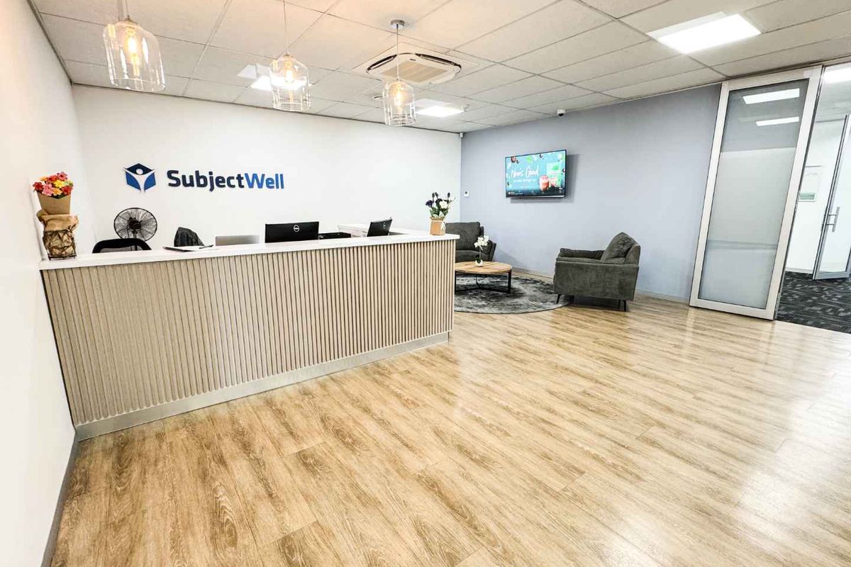 Modern office reception area featuring a custom reception desk, wood-look laminate flooring, suspended ceiling with integrated lighting, and a glass partition entrance.