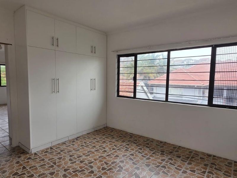 A bright, empty bedroom featuring built-in cupboards, tiled flooring, and large windows with security bars, offering a clean and functional space.