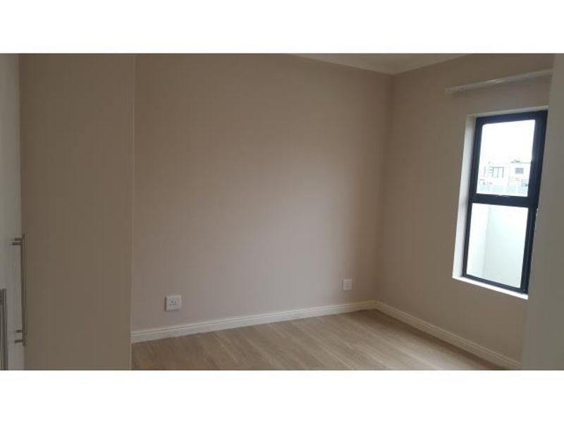 A clean, empty bedroom featuring neutral wall tones, modern laminate flooring, and a built-in cupboard with a sleek aluminium-framed window.