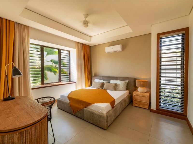 A well-maintained bedroom featuring a tray ceiling, split-system air conditioning, large windows with wooden frames, and neutral tiled flooring.