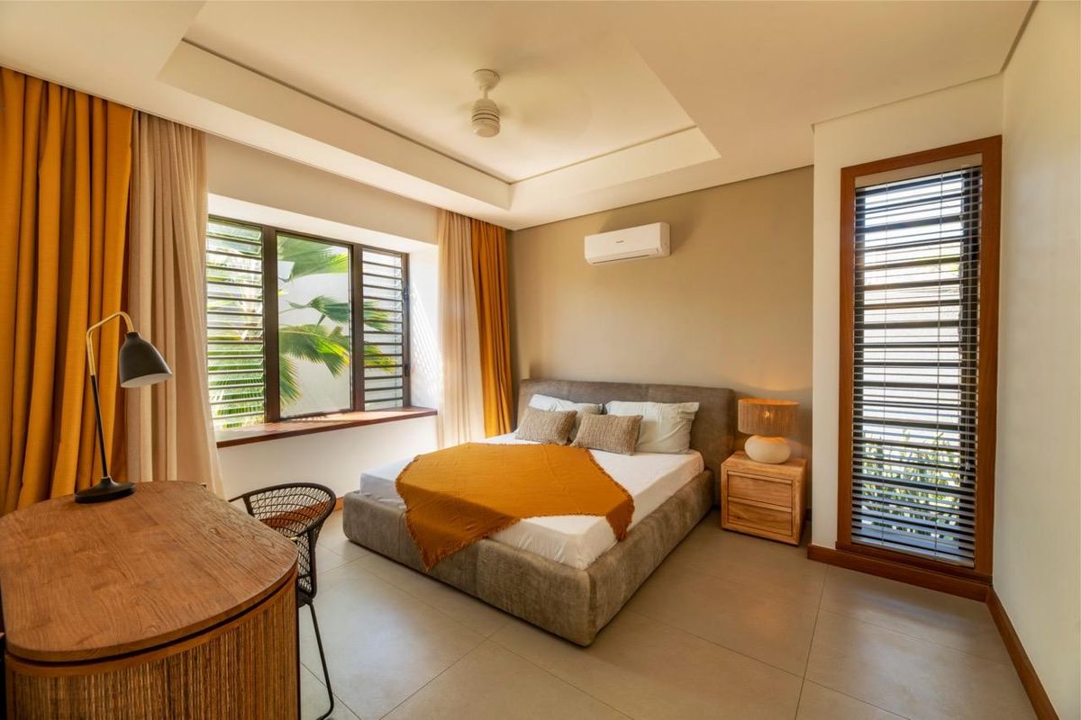 A well-maintained bedroom featuring a tray ceiling, split-system air conditioning, large windows with wooden frames, and neutral tiled flooring.
