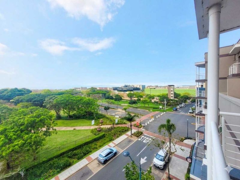 Elevated view from a balcony overlooking a landscaped park, street intersection, and surrounding urban development.