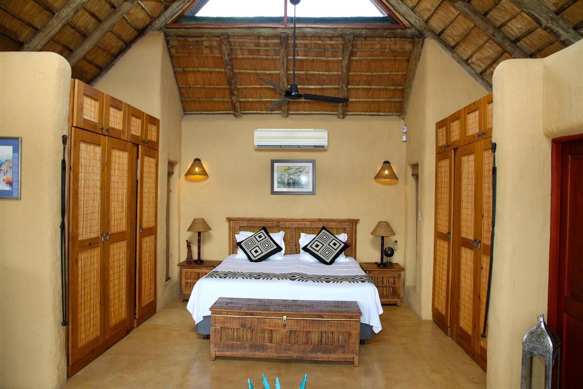 A spacious bedroom featuring a high vaulted thatched ceiling, exposed timber beams, built-in wooden cabinetry, and a wall-mounted air conditioning unit.