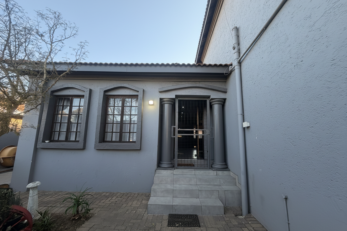 Rear exterior view featuring a paved patio, tiled entrance steps with decorative columns, and grid-style windows on a grey rendered facade.