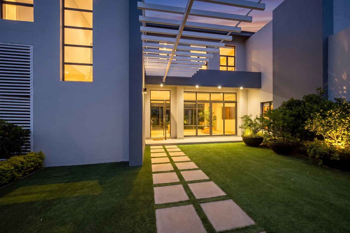 Modern rear exterior featuring a paved walkway, pergola structure, and illuminated glass doors leading into the home.