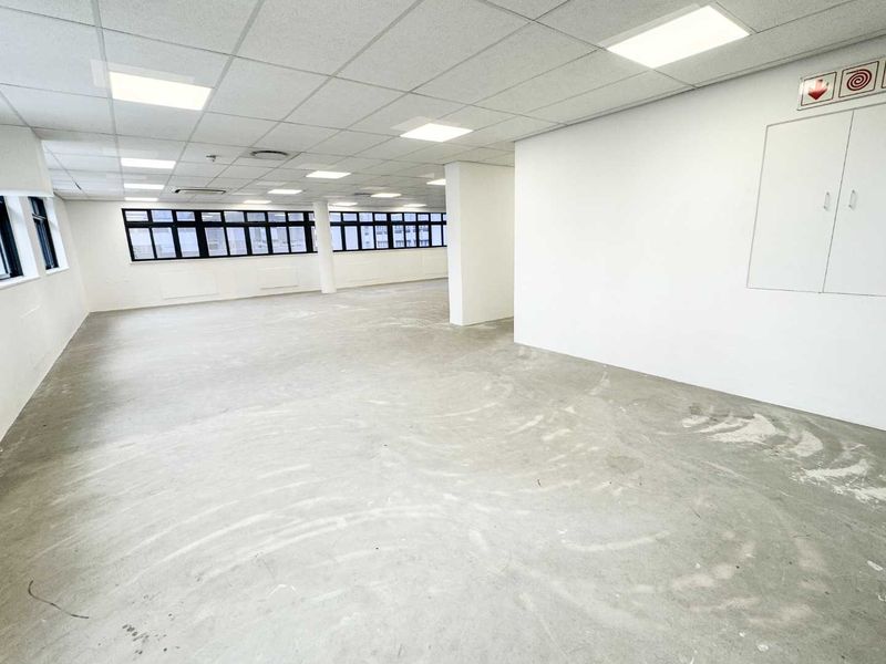A spacious, open-plan office area featuring a suspended ceiling with recessed lighting, large windows for natural light, and an unfinished concrete floor.
