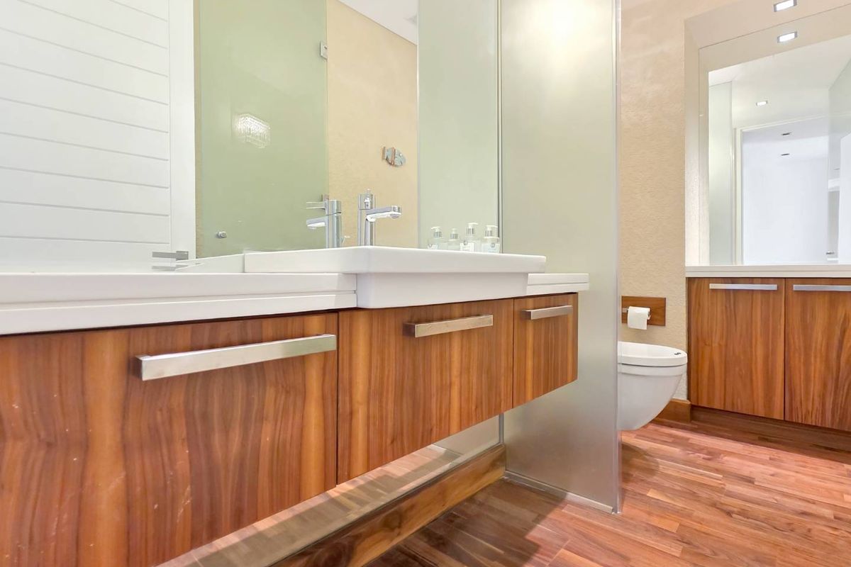 Modern bathroom featuring a floating timber vanity, integrated basin, wall-mounted toilet, and a sleek frosted glass partition with wood-look flooring.