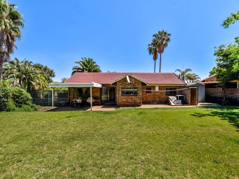 Rear view of a brick house featuring a tiled roof, a covered patio area, and a spacious garden with established palm trees.