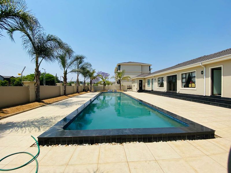 A well-maintained outdoor pool area featuring a long rectangular swimming pool with dark stone coping, surrounded by paved decking and a perimeter boundary wall.