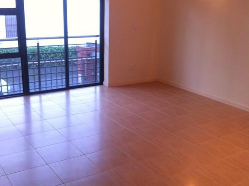 An empty living room featuring neutral tiled flooring, a wall-mounted light fixture, and a large sliding glass door providing access to the balcony.
