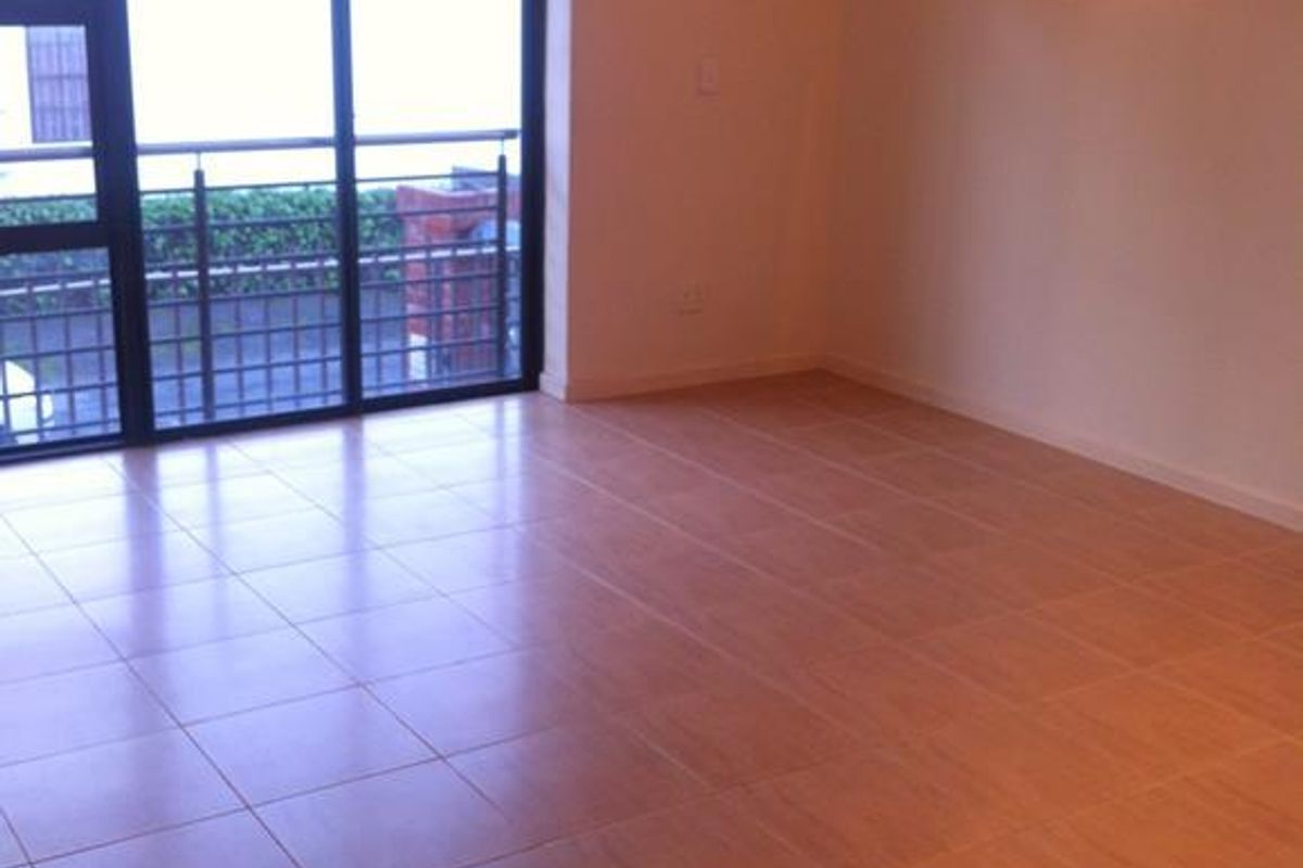 An empty living room featuring neutral tiled flooring, a wall-mounted light fixture, and a large sliding glass door providing access to the balcony.