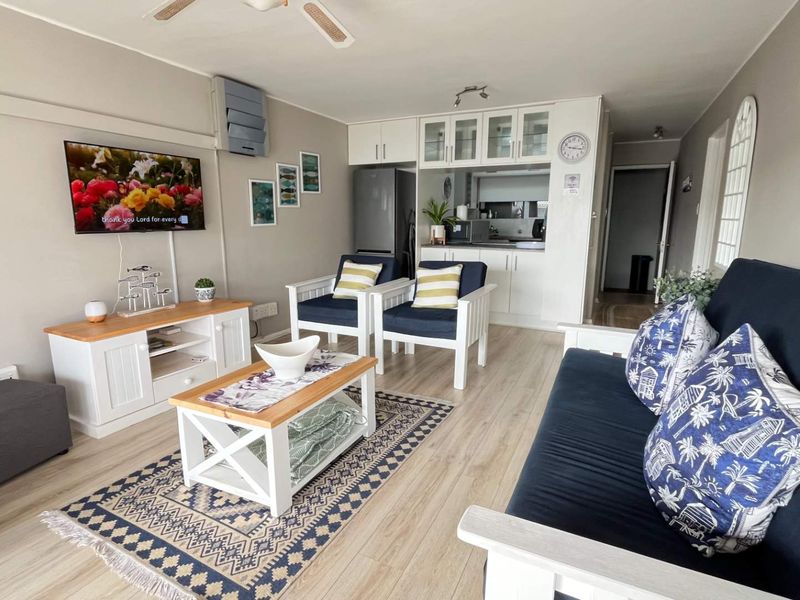 An open-plan living area featuring light laminate flooring, a ceiling fan, and a compact kitchen with white cabinetry and glass-fronted display units.