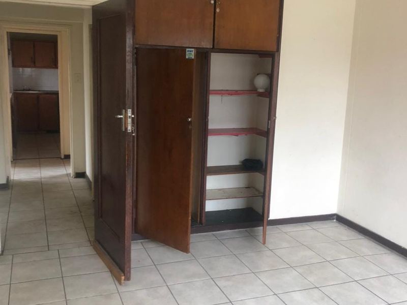 An empty bedroom featuring built-in wooden cupboards and ceramic floor tiles, with a view through to the kitchen area.
