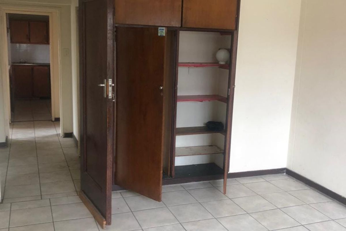 An empty bedroom featuring built-in wooden cupboards and ceramic floor tiles, with a view through to the kitchen area.