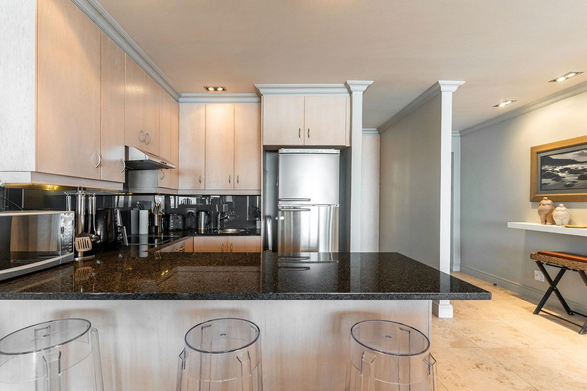 Modern kitchen featuring light wood cabinetry, dark granite countertops, a breakfast bar, and integrated stainless steel appliances, flowing into an adjacent living area.