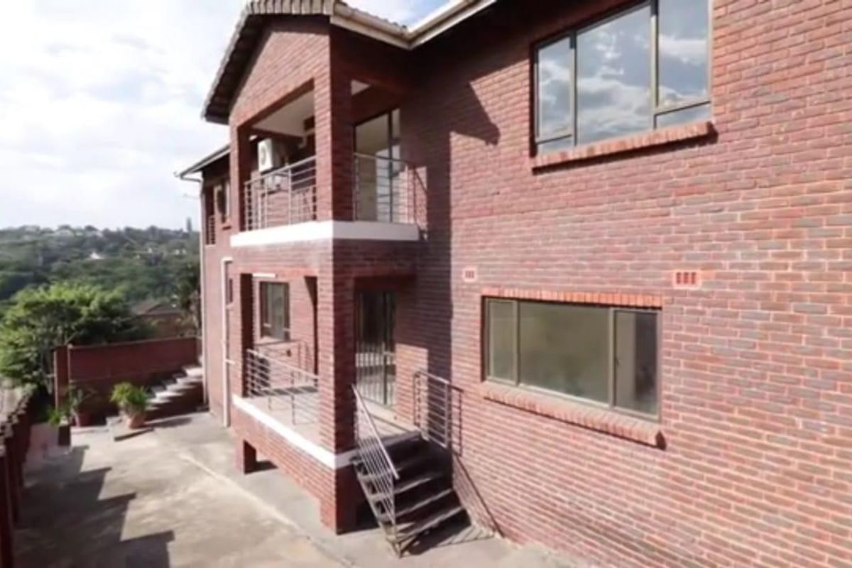 A rear view of a double-story face brick house featuring balconies with metal railings, aluminum windows, and a paved outdoor area.