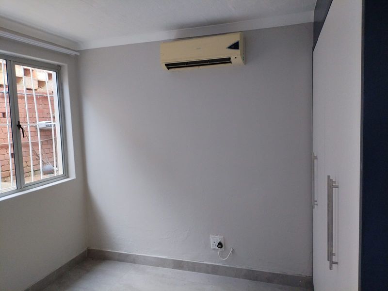 A clean, empty bedroom featuring tiled flooring, built-in cupboards, and an installed air conditioning unit with security bars on the window.