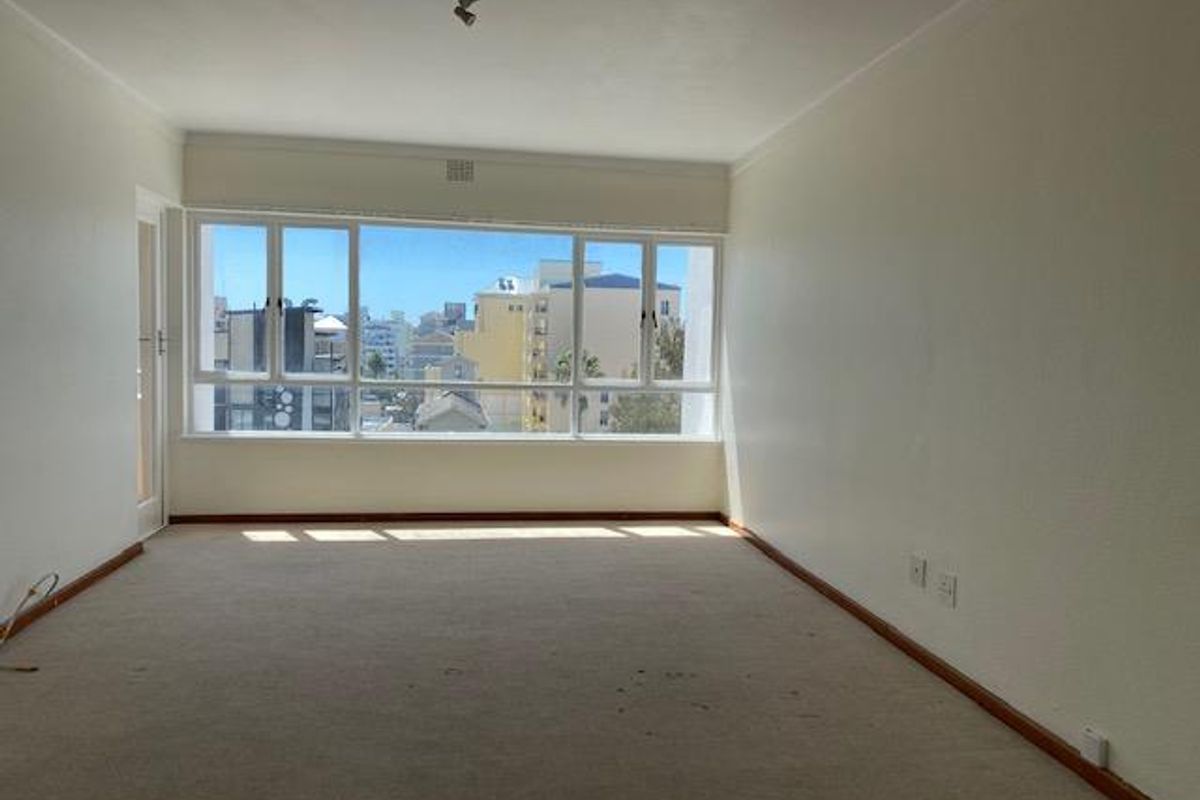 An empty living room featuring large windows with urban views, neutral walls, and carpeted flooring, offering a blank canvas for renovation.