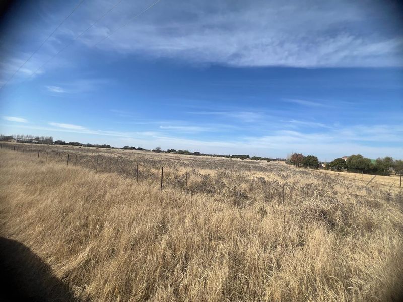 A wide, open residential vacant land plot featuring natural dry grass and perimeter wire fencing under a clear blue sky.