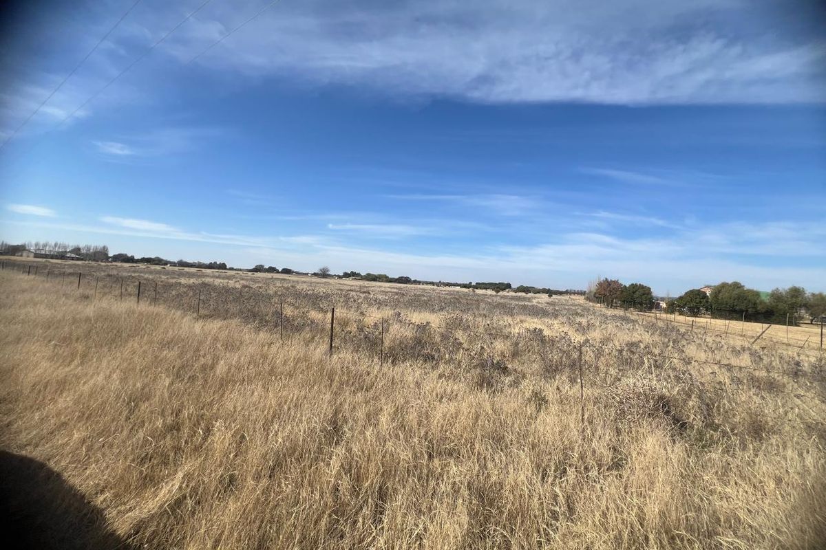 A wide, open residential vacant land plot featuring natural dry grass and perimeter wire fencing under a clear blue sky.