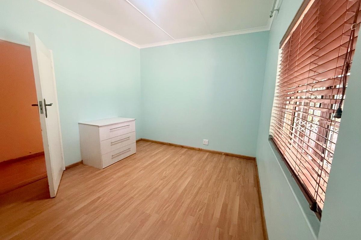 A bright, empty bedroom featuring light-toned laminate flooring, pale blue walls, and fitted venetian blinds.