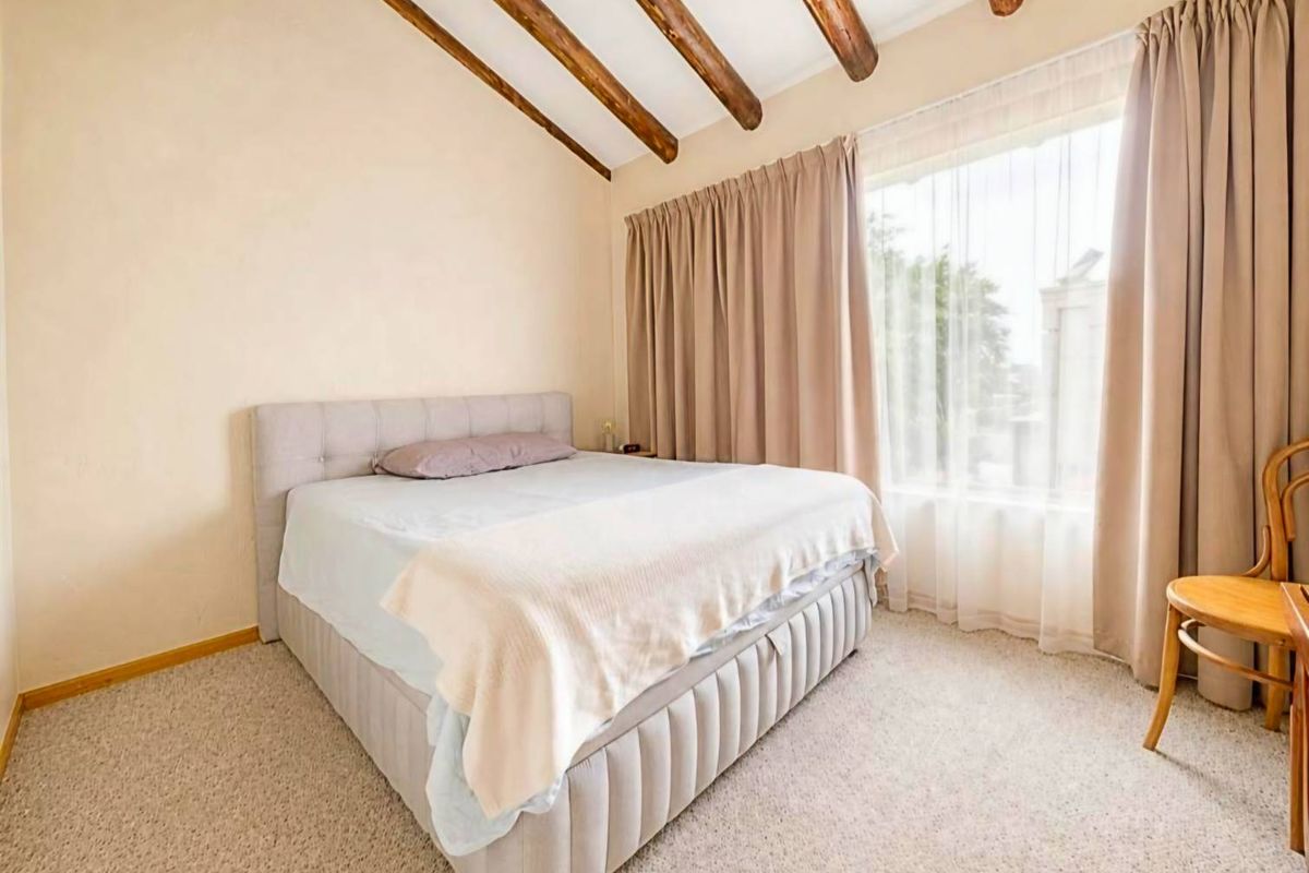 A cozy bedroom featuring a vaulted ceiling with exposed timber beams, neutral carpeting, and a large window allowing for ample natural light.