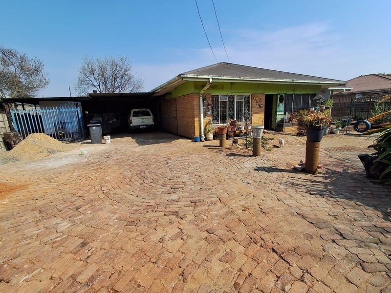 Front exterior view of a single-story house featuring a brick paved driveway, attached carport, and security gates.