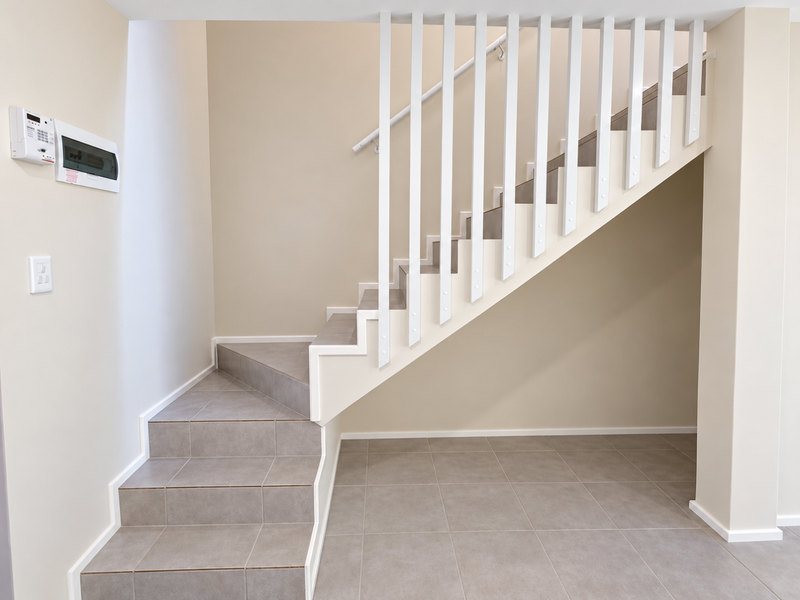 A clean, modern hallway featuring a tiled staircase with a white balustrade, neutral wall tones, and integrated electrical and security fixtures.