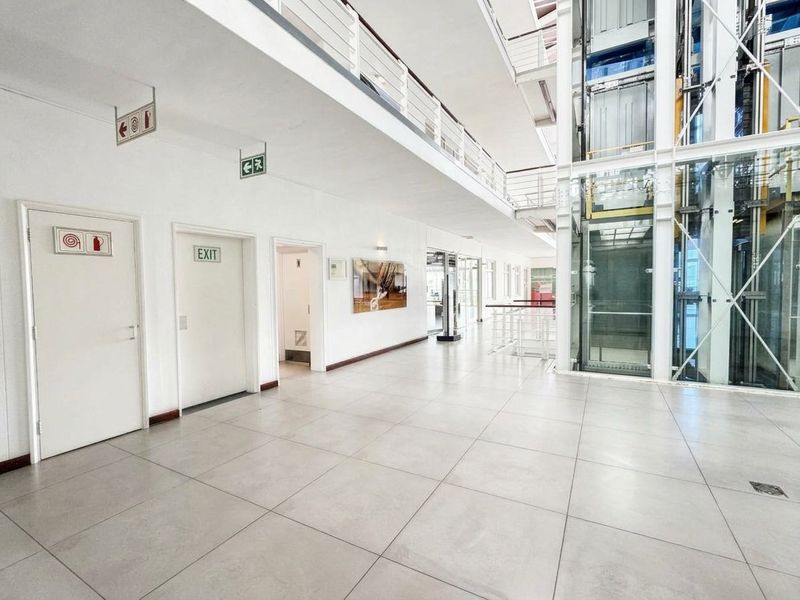 Spacious commercial hallway featuring a modern glass elevator, tiled flooring, and a mezzanine level with metal balustrades.