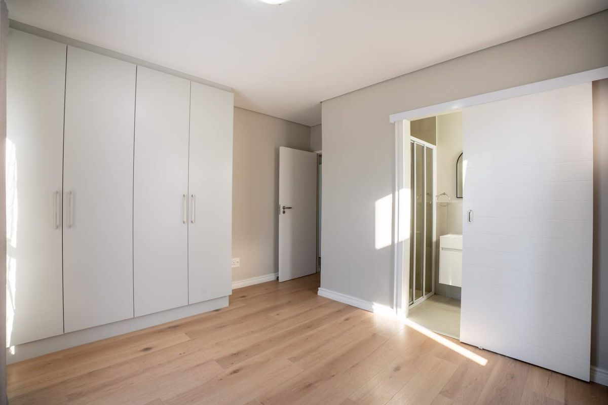 A bright, modern bedroom featuring light wood-look laminate flooring, ample built-in cupboard space, and a sliding door leading to an en-suite bathroom.
