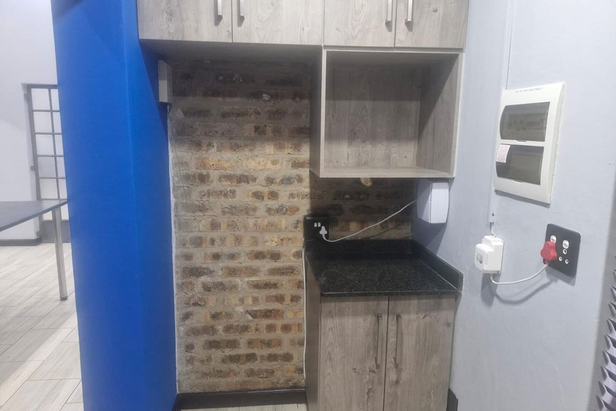 A compact kitchenette area featuring wood-look cabinetry, granite countertop, and an exposed brick feature wall within an office space.