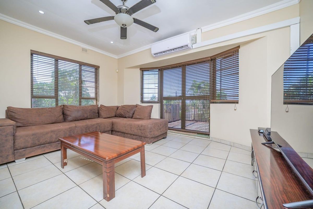 A spacious living room featuring tiled flooring, a ceiling fan, and a wall-mounted split-system air conditioner, with large windows fitted with venetian blinds.